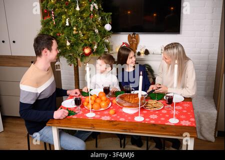 Glückliche Familie unterhält sich am Weihnachtstisch, Eltern und Kinder lächeln während des festlichen Feiertagsessens in der Nähe eines leuchtenden Weihnachtsbaums, gemütliche Winterabendfeier, Konzept des häuslichen Lebensstils Stockfoto