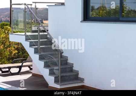 Eine zeitgenössische Granittreppe mit eleganten Metall- und Glasgeländern steigt neben einer weißen Stuckwand auf Stockfoto
