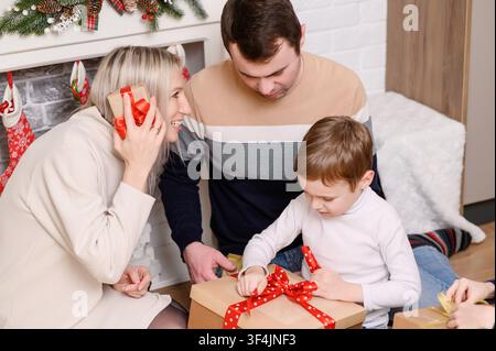 Glückliche Familie sitzt auf dem Boden in der Nähe eines geschmückten Kamins, um Weihnachtsgeschenke auszupacken. Junge zieht an der roten Schleife einer großen Bastelschachtel, während die Mutter lächelt und ein kleineres Geschenk schüttelt. Konzept für Winterfeiern Stockfoto