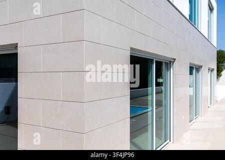 Eine zeitgenössische architektonische Fassade zeigt großformatige hellbeige Steinfliesen und weitläufige Glastüren, die einen blauen Swimmingpool reflektieren Stockfoto