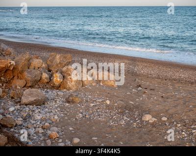 Große Felsbrocken und Steine an einem sandigen Kiesstrand neben blauen Meereswellen unter einem klaren Himmel bei sanftem Licht. Stockfoto
