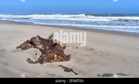 Ein stark verrostetes Maschinenteil aus dem Schiffswrack der Benguela liegt am sandigen Ufer, während sich im Hintergrund die Wellen des Atlantischen Ozeans brechen, in Namibia. Stockfoto