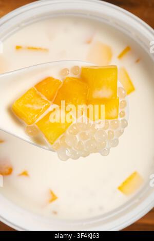 Löffel gefüllt mit Mangowürfeln und Sago über einer Schüssel mit cremigem Mango-Sago-Dessert Stockfoto