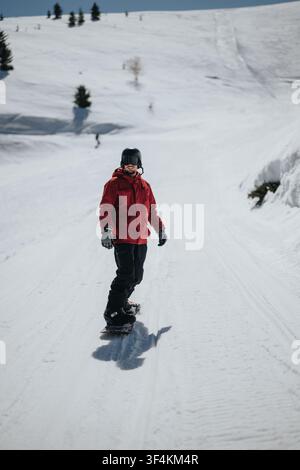Snowboarder in roter Jacke fährt einen schneebedeckten Bergabhang hinunter mit Helm und Skibrille Stockfoto