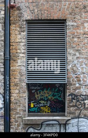 Nahaufnahme einer Ziegelmauer und eines Fensters, das mit Metalljalousien und urbaner Graffiti-Markierung im Zentrum von Dublin, Irland, bedeckt ist. Stockfoto