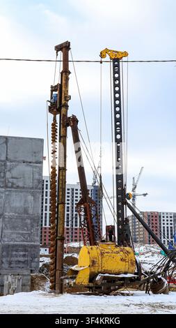 Eine große Bohrplattform ist auf einer Baustelle für ein Haus mitten im Schnee aufgestellt, um Löcher für Pfahlfundamente zu bohren. In der Nähe befinden sich Kräne und unfertige B Stockfoto