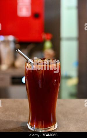 Ein erfrischendes Glas mit amerikanischem Eiskaffee, gefüllt mit Eiswürfeln und einem wiederverwendbaren Metallstrohhalm, steht auf einer hölzernen Theke in einem modernen Café. Perfekt für Kaffeeshop-Menüs, Sommergetränkekonzepte und Lifestyle-Visuals. Stockfoto