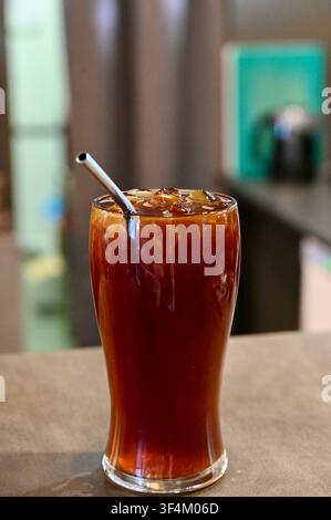 Ein erfrischendes Glas mit amerikanischem Eiskaffee, gefüllt mit Eiswürfeln und einem wiederverwendbaren Metallstrohhalm, steht auf einer hölzernen Theke in einem modernen Café. Perfekt für Kaffeeshop-Menüs, Sommergetränkekonzepte und Lifestyle-Visuals. Stockfoto