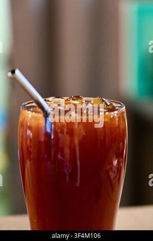 Eine detaillierte Nahaufnahme eines Glases gefüllt mit kaltem Americano-Kaffee, Eiswürfeln und einem wiederverwendbaren Metallstrohhalm in einer modernen Café-Umgebung. Perfekt für Kaffeemenüs, Sommererfrischungskampagnen und Café-Promotionsdesigns. Stockfoto