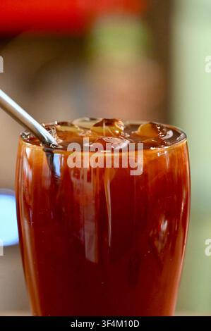 Eine detaillierte Nahaufnahme eines Glases gefüllt mit kaltem Americano-Kaffee, Eiswürfeln und einem wiederverwendbaren Metallstrohhalm in einer modernen Café-Umgebung. Perfekt für Kaffeemenüs, Sommererfrischungskampagnen und Café-Promotionsdesigns. Stockfoto