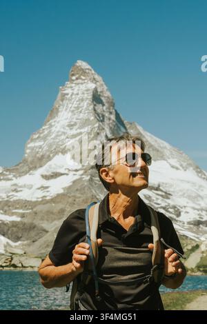 Mann lächelt, trägt Sonnenbrille, beim Backpacking vor einem schneebedeckten Matterhorn-Gipfel und einem See Stockfoto