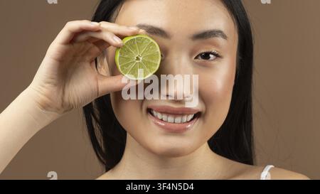 Lächelnde asiatische Frau hält Limettenstück vor dem Auge, Nahaufnahme, Konzept für einen gesunden Lebensstil Stockfoto