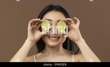 Lächelnde asiatische Frau hält Limettenstücke über die Augen, fröhlicher Ausdruck, Kücheneinrichtung Stockfoto