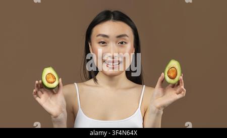 Lächelnde junge asiatische Frau hält Avocado-Hälften, gesundes Lebensmittelkonzept Stockfoto