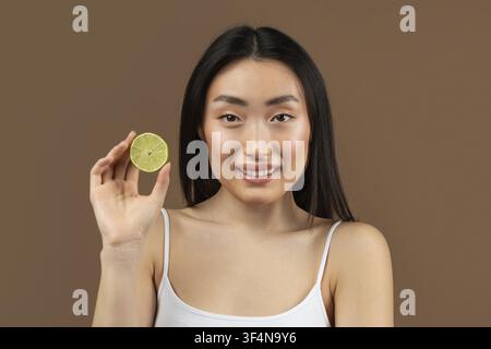 Lächelnde asiatische Frau hält Limette mit fröhlichem Ausdruck, Konzept für einen gesunden Lebensstil Stockfoto