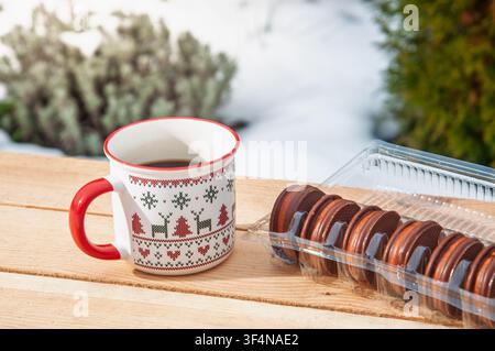 Eine festliche Tasse gefüllt mit dunklem Kaffee steht neben einer Packung Schokoladen-Sandwich-Keksen auf einer rustikalen Holzoberfläche mit schneebedecktem Hintergrund Stockfoto