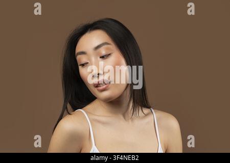 Junge asiatische Frau posiert mit nachdenklichem Ausdruck, brauner Hintergrund Stockfoto