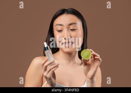 Glückliche junge asiatische Frau hält Serum und Limettenscheiben in der Hand, Hautpflegekonzept Stockfoto