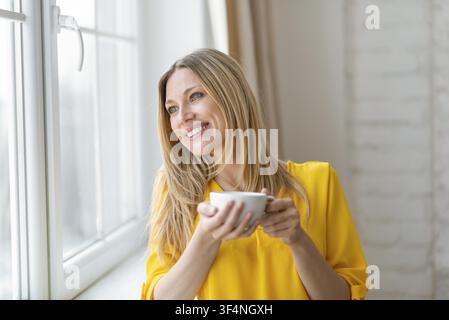 Fröhliche junge Frau, die eine Tasse hält und am Fenster lächelt, Innenstudio-Lebensstilkonzept Stockfoto