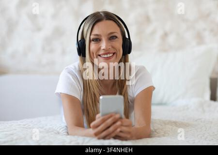 Zufriedene junge Frau hört Musik mit Kopfhörern im Bett, entspannt zu Hause Stockfoto