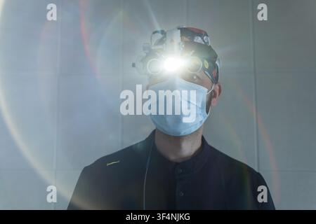 Professioneller Chirurg trägt Gesichtsmaske und chirurgische Lupen mit heller Kopflampe, die direkt leuchtet und Lichtreflexe erzeugt. Nahaufnahme-Porträt eines medizinischen SP Stockfoto