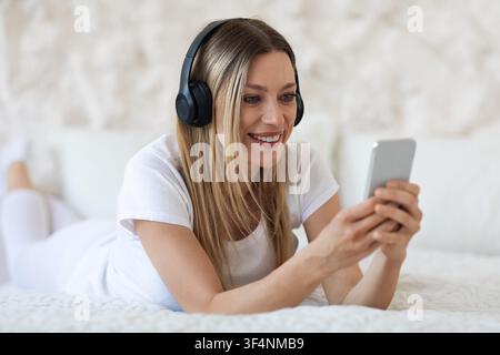 Lächelnde junge Frau hört Musik mit Kopfhörern und benutzt ein Smartphone zu Hause Stockfoto