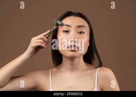 Junge asiatische Frau verwendet einen Jade-Roller für ihre Hautpflege-Routine, gesundes Strahlen, Konzept für den Heim-Spa Stockfoto