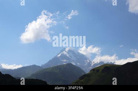 Panoramatische Ansicht des schneebedeckten Berges Kazbegi unter einem weiten blauen Himmel mit leichten Wolken und grünen Bergsilhouetten im Vordergrund, Georgien Stockfoto