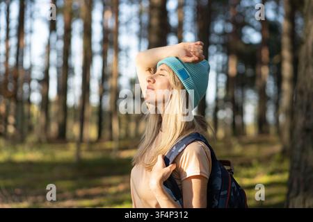Fröhliche Reisende blinkend und genießt Sonnenlicht, während sie mit dem Rucksack durch den Wald spaziert Stockfoto