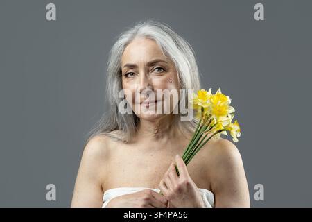 Reife Frau hält gelbe Narzissen mit sanftem Ausdruck, Studio-Porträt, Schönheitskonzept Stockfoto