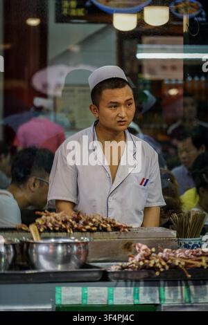 UNBEKANNT, China - 25. August 2017: Ein junger Mann, der einen weißen Kochkittel mit blauem Saum und eine kleine blaue Kappe trägt, steht hinter einem Imbissstand Stockfoto
