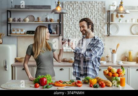 Glücklicher kaukasischer Mann und Frau stoßen mit Rotweingläsern in einer modernen Küche an, fröhliche Atmosphäre Stockfoto