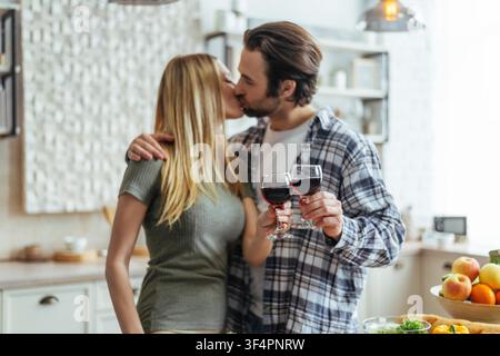 Glückliches junges Paar küsst sich in der Küche und hält Gläser mit Rotwein, romantischer Moment Stockfoto