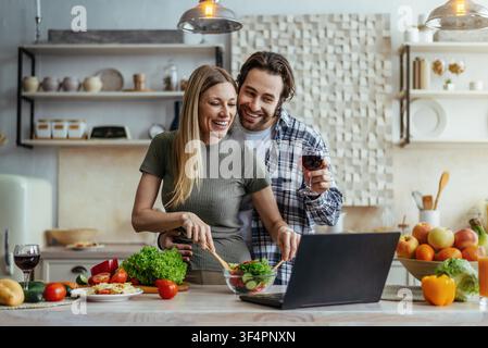 Glückliches junges Paar kocht zusammen in der Küche, bereitet einen gesunden Salat mit Laptop zu, organisches Konzept Stockfoto