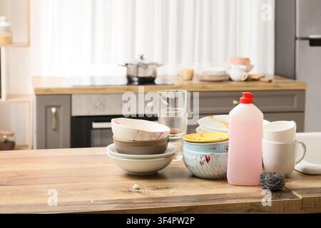 Stapel schmutziger Teller und Gläser auf einer hölzernen Küchentheke mit Spülmittel und einem Schwamm, eine Flasche rosa Spülmittel mit rotem Deckel und einem Stahlwischer Stockfoto