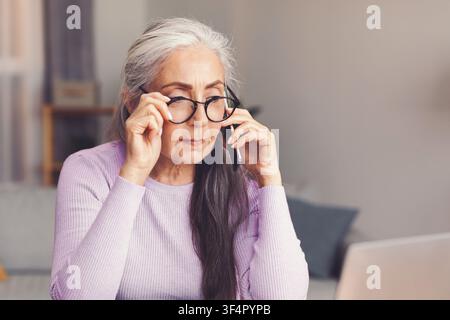 Ältere Frau mit grauen Haaren, die einen Laptop und ein Telefon im Heimbüro benutzt und sich auf das Gespräch konzentriert Stockfoto