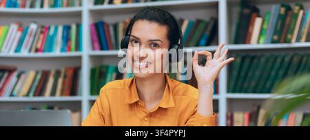 Lächelnde junge hispanische Frau mit Kopfhörern, die in der Bibliothek an einem Laptop arbeitet, Bildungsbegriff Stockfoto