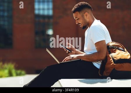 Junger schwarzer Mann, der einen Laptop und ein Smartphone benutzt, während er im Freien sitzt, Geschäftskonzept Stockfoto