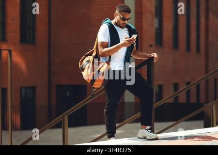 Afroamerikanischer Mann überprüft sein Smartphone, während er auf Treppen steht, lässige Außeneinrichtung Stockfoto