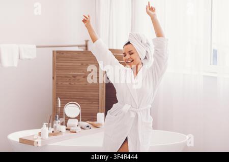 Glückliche Frau genießt ihre Morgenroutine in weißem Bademantel und Handtuch, Badezimmer, Wellness-Konzept Stockfoto