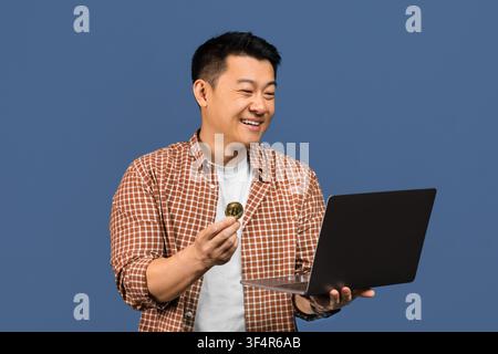 Zufriedener Asiate, der Bitcoin hält, während er einen Laptop benutzt, Geschäftskonzept Stockfoto