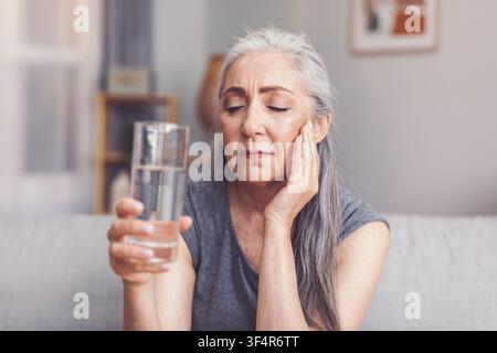 Nachdenkliche ältere Frau, die ein Glas Wasser zu Hause hält, konzentriert auf das Konzept von Gesundheit und Wohlbefinden Stockfoto