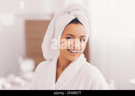Lächelnde junge Frau mit Handtuch auf dem Kopf in einem weißen Bademantel, Hautpflegeroutine zu Hause Stockfoto