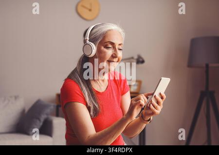 Zufriedene ältere kaukasische Frau, die zu Hause Musik hört und ein Smartphone benutzt, Lebensstilkonzept Stockfoto