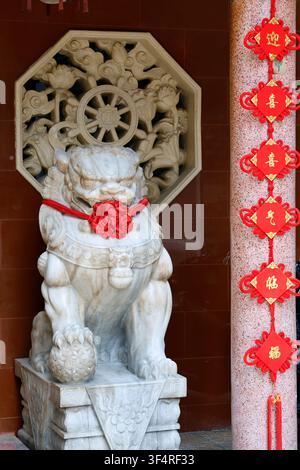 Van Phat Pagode, oder Tempel der 1.000 Buddhas. Eine mit einem roten Band geschmückte Marmorwächterlöwenstatue steht vor einem Wandrelief, das th Stockfoto