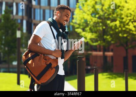 Lächelnder schwarzer männlicher Student verwendet Smartphone beim Gehen im Freien, Gewichtsabnahme-Konzept Stockfoto