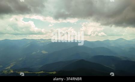 Sanfte Sonnenstrahlen brechen durch dicke Wolken und beleuchten entfernte Kämme, während Schichten aus blauen Bergen im Dunst verschwinden – eine ruhige Hochland-Szene voller Luftigkeit Stockfoto
