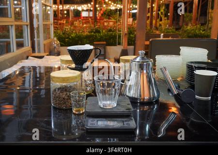 Manuelle Pour-Over-Kaffeebereitstellungs-Einrichtung mit Kaffeetropfer, Wasserkocher, Kaffeebohnen und Waage auf einer Café-Theke bei Nacht. Stockfoto
