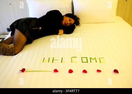 Eine Frau entspannt sich auf einem Hotelbett, wo das Wort "Willkommen" aus Rosenblättern geschrieben ist. Pattaya, Thailand. Stockfoto