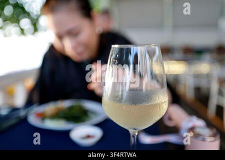 Ein Glas gekühlter Weißwein wird auf einem Restauranttisch gezeigt, mit einer verschwommenen Figur im Hintergrund. Stockfoto